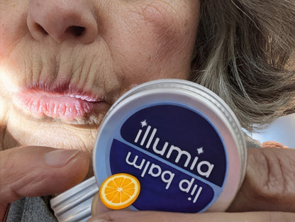 Person holding a container of Illumia lip balm with an orange slice for the orange-aid variant.